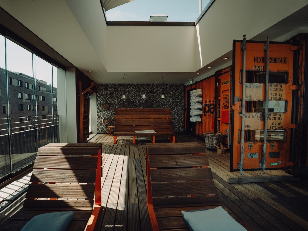 Modern sauna recreaation area with wooden deck chairs, bright windows, and an industrial-style wall featuring colorful shipping containers.