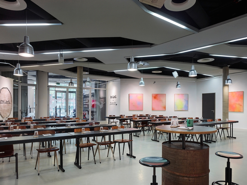 Modern event space featuring minimalist design, wooden tables, colorful abstract paintings, and industrial pendant lighting.
