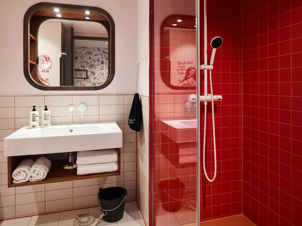 Modern bathroom featuring a red tiled shower, white sink, stylish mirror, and neatly arranged towels.