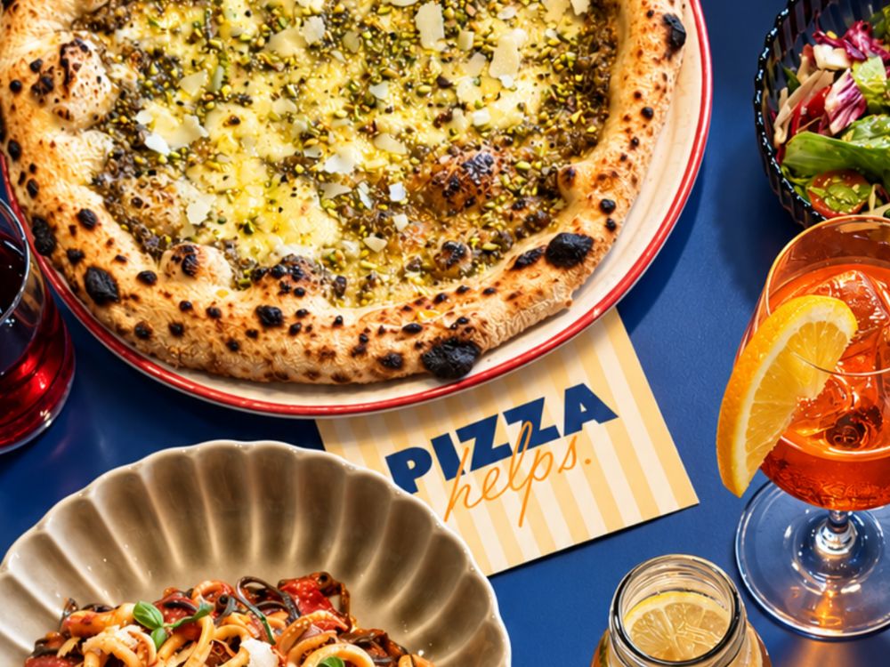 Flat lay of a Neapolitan pizza with pesto, a bowl of pasta, a side salad, and an Aperol Spritz on a blue table. 