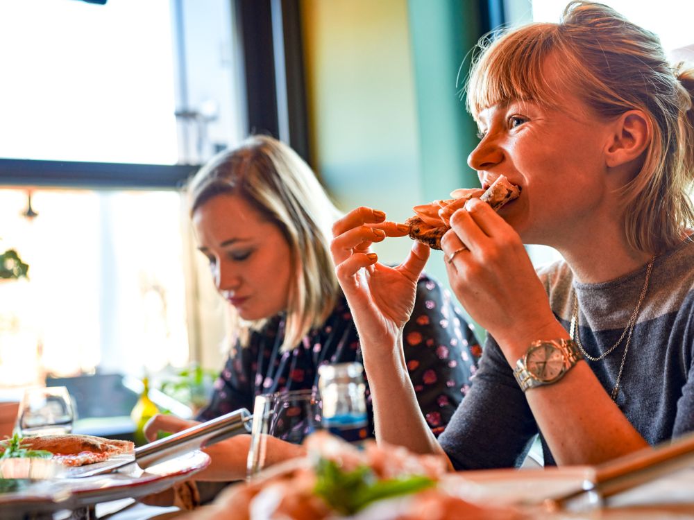 A cheerful blonde woman smiling while taking a bite of a fresh slice of pizza at a restaurant.