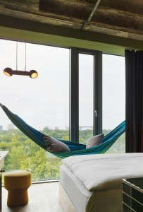 A hotel room featuring a blue hammock by a large window, a plush bed, and an inviting yellow stool.