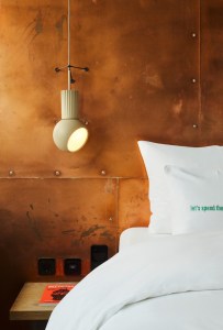 A bed with white linens, a decorative pillow, and a modern pendant light against a warm copper wall. A book lies on the bedside table.