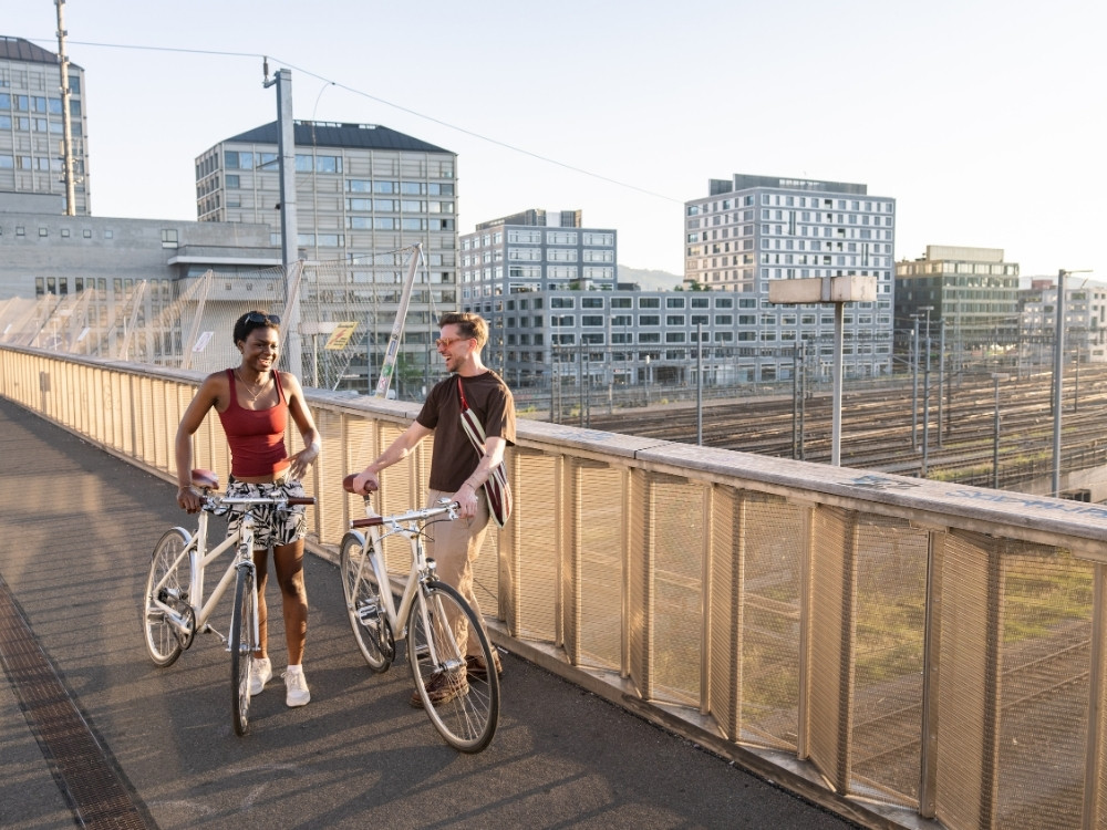 Two people walking bicycles on a city bridge, smiling and chatting, with modern buildings and train tracks in the background.