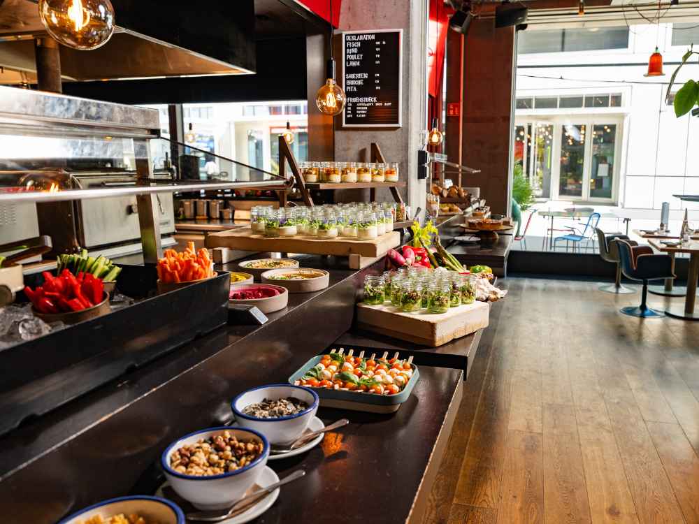 A modern and colorful hotel buffet counter featuring fresh vegetable sticks, gourmet small jars with appetizers, nuts, and Caprese skewers next to an open kitchen.