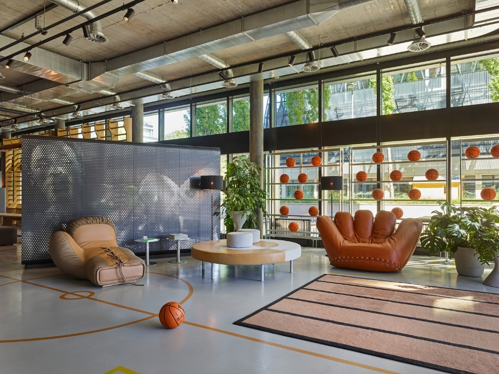 A modern lobby area featuring unique furniture, a basketball-themed decor, large windows, and plants amidst a vibrant atmosphere.