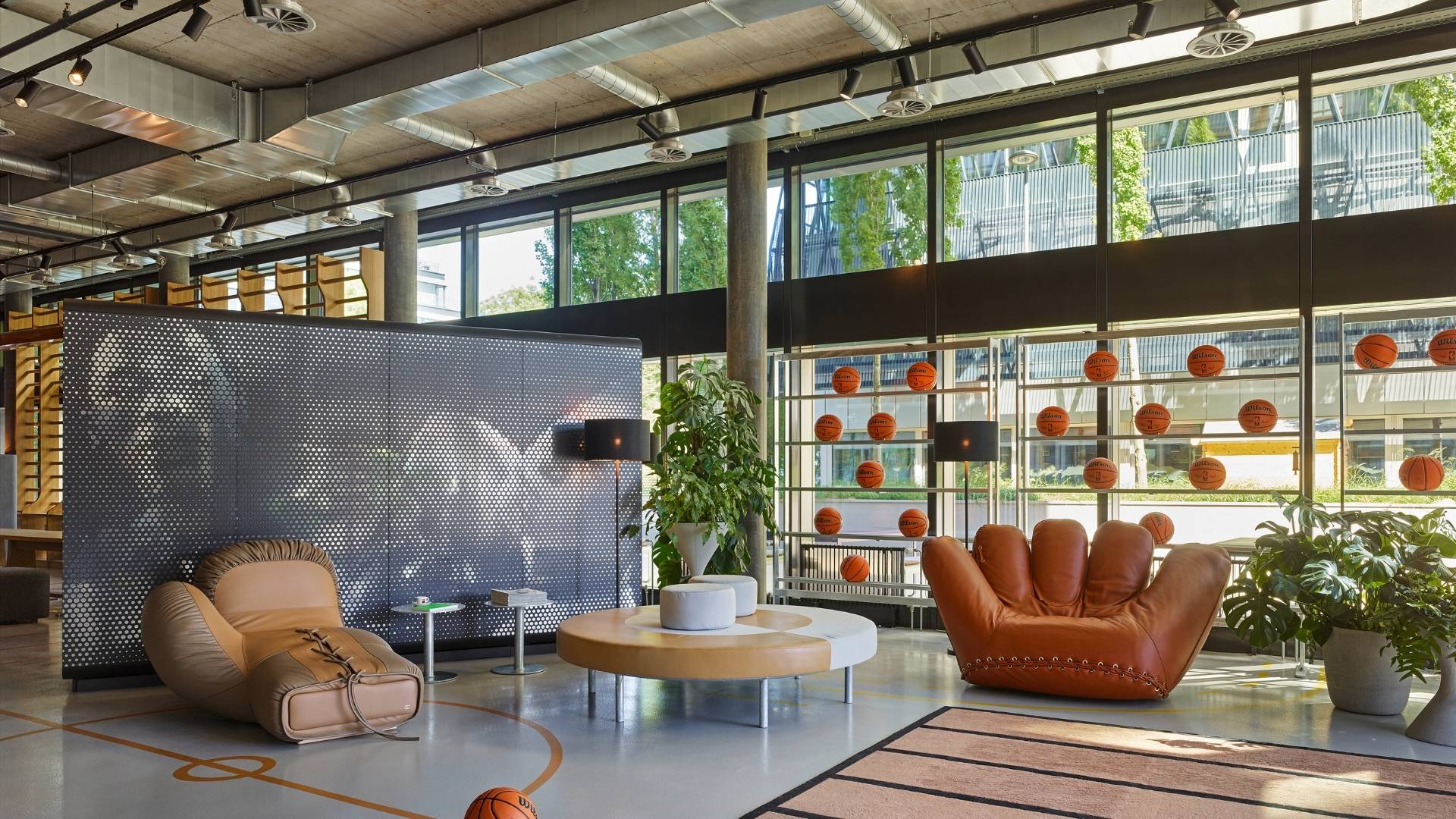A modern lobby area featuring unique furniture, a basketball-themed decor, large windows, and plants amidst a vibrant atmosphere.