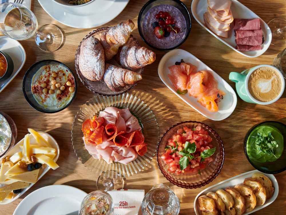 Overhead view of a vibrant breakfast table layout featuring fresh croissants, smoked salmon, cold cuts, sliced cheese, hummus, tomato salsa, and hot cappuccino.
