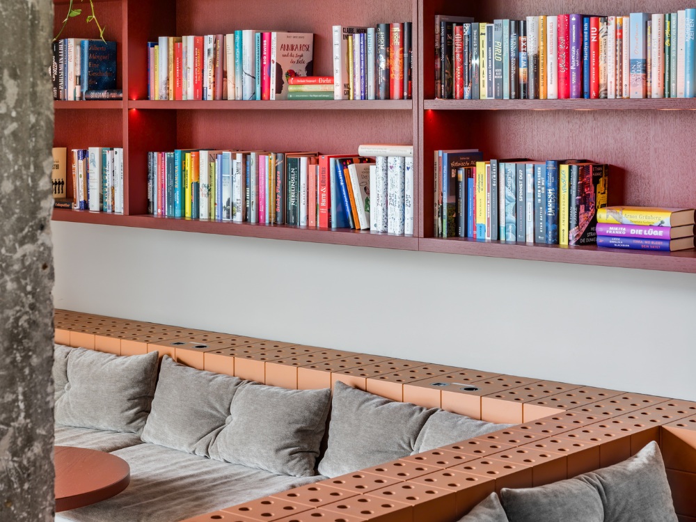 Cosy café with a padded gray sofa and soft cushions. Above, a bookshelf filled with colorful books creates a warm and inviting atmosphere.