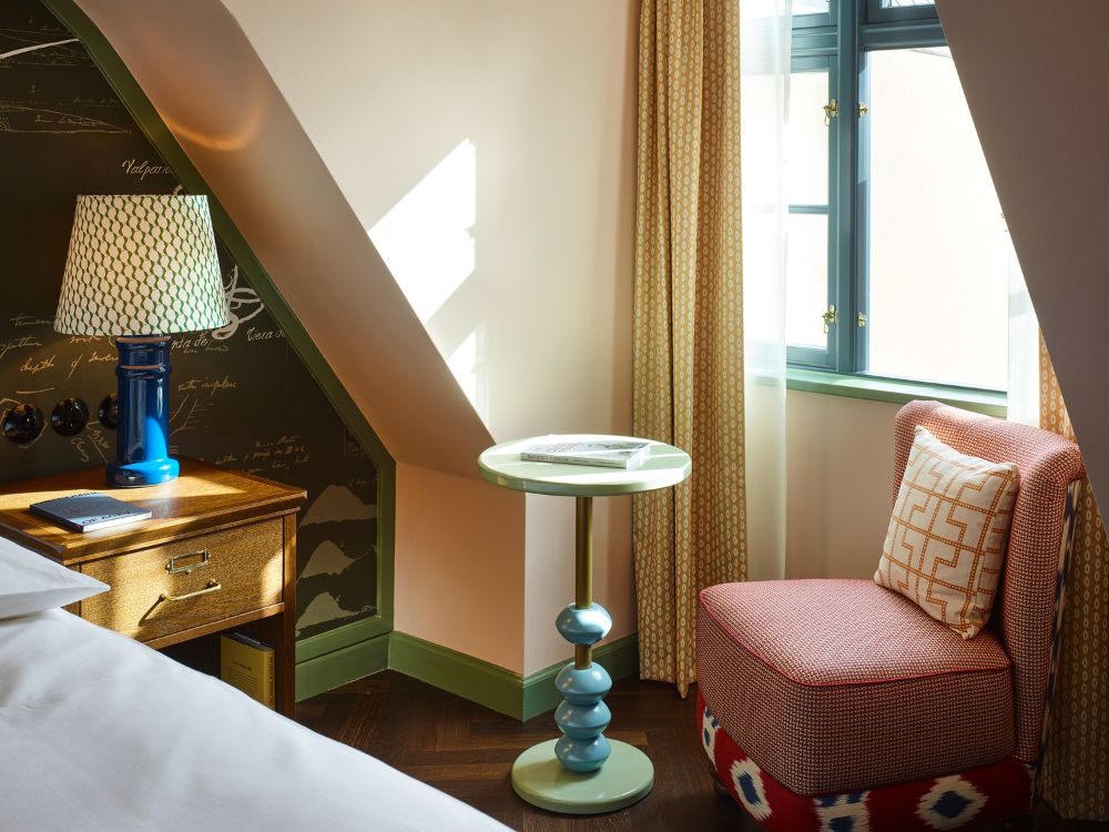Cozy room corner with a red-patterned chair, geometric cushion, round turquoise table, and a decorative lamp on a wooden side table by a window.