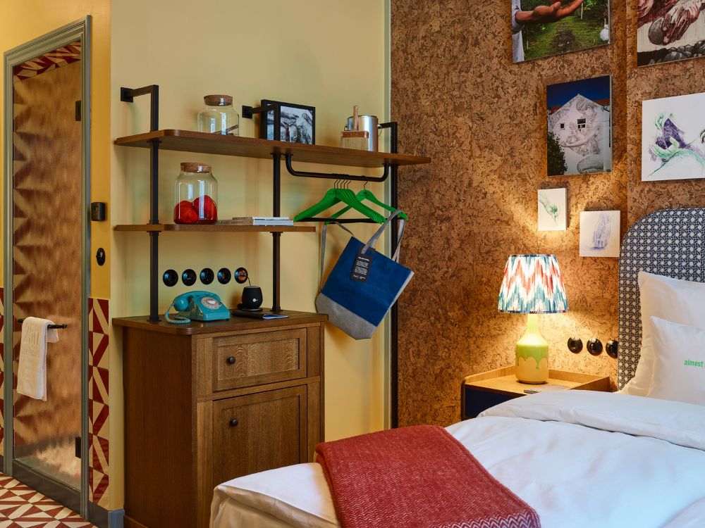 Cozy hotel room with eclectic decor: a cork wall with art, colorful lamp, wooden shelves with retro items, and a bed with a red blanket. Inviting ambiance.