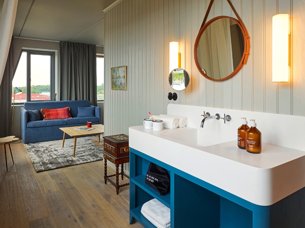 Hotel room with a blue sofa topped by red and blue pillows, wooden coffee table, and a rug. Adjacent is a sleek sink with round mirror and warm lighting.