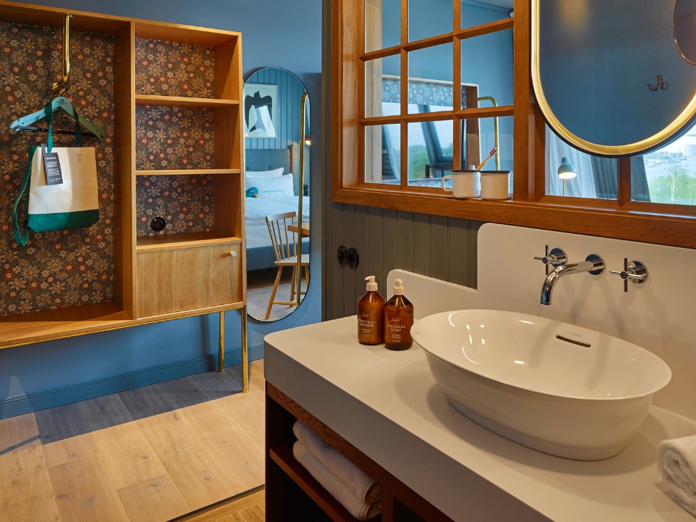 Bathroom with a white basin and chrome faucet. Wooden shelving, patterned wallpaper, and toiletries add a cosy, upscale touch.