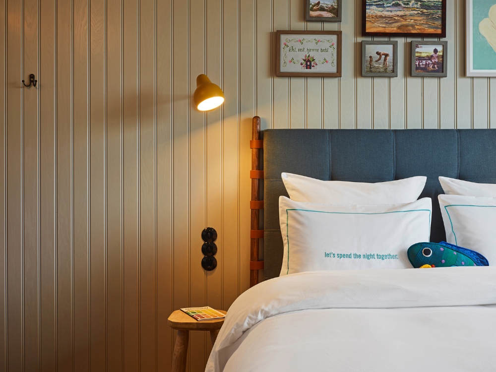 Bedroom with a modern design, featuring a blue upholstered headboard, pillows saying “let’s spend the night together,” wall art, and a wooden nightstand.