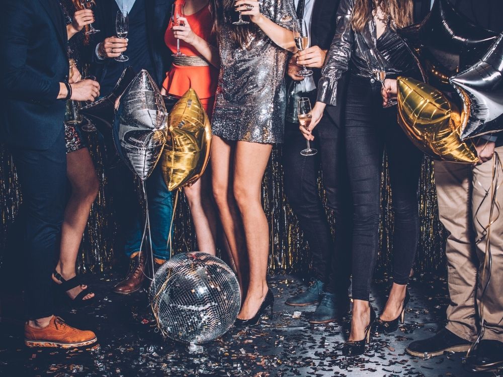 A group of stylishly dressed individuals celebrating with champagne and balloons, standing around a sparkling disco ball.