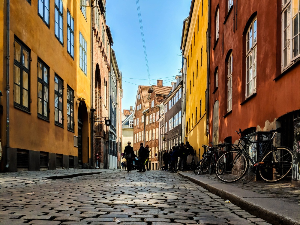 Narrow cobblestone street flanked by colorful buildings in Copenhagen; people gather and bicycles are parked along the vibrant walls under a clear sky.