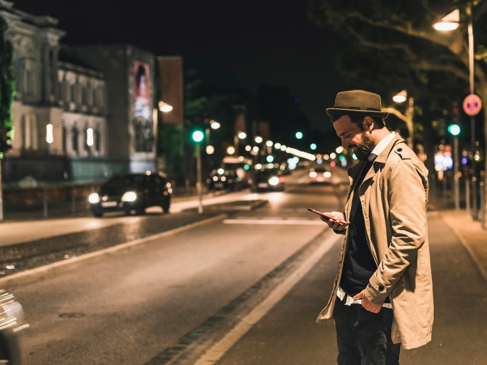 A man in a tan trench coat and hat checks his smartphone while standing on a dimly lit street at night in Frankfurt, surrounded by passing cars.