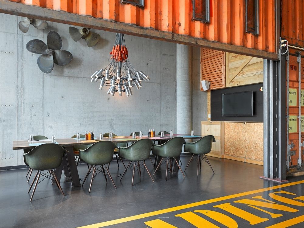 Meeting room in a shipping container featuring a long table, green chairs, industrial decor, and a unique chandelier made from pipes and light bulbs.