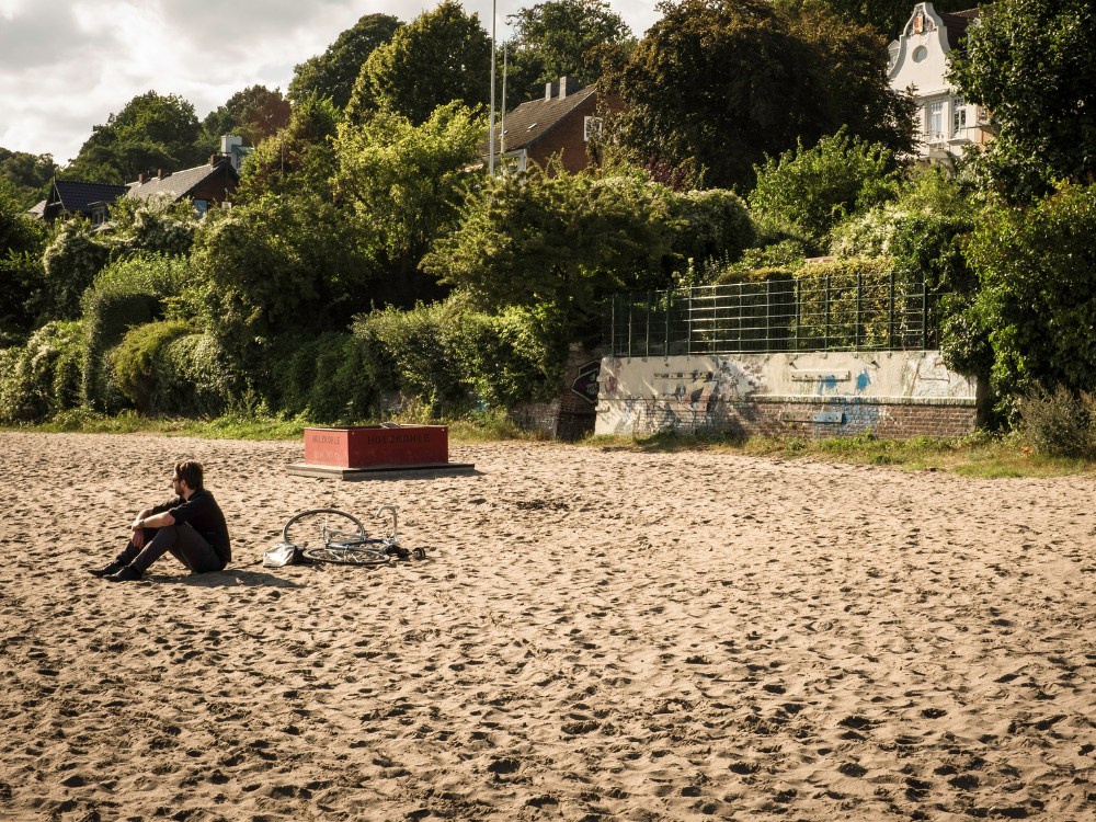 A lone person sitting on a sandy beach, with a bicycle nearby and green trees in the background, evoking a peaceful, reflective moment.