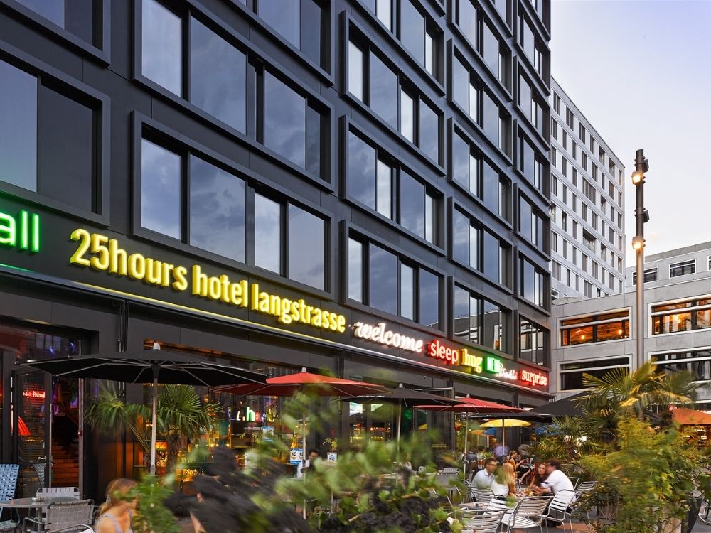 Modern hotel exterior at dusk with reflective windows and neon signs reading "25hours hotel langstrasse." Outdoor seating area with people and plants.