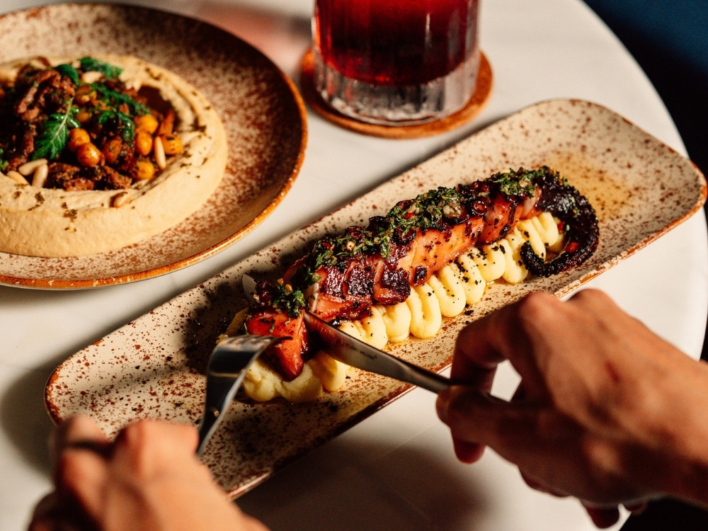 A hand holds a fork and knife, preparing to cut into grilled octopus atop creamy mashed potatoes, with a plate of hummus in the background.