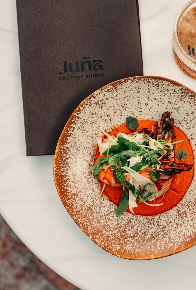 A gourmet dish of grilled shrimp atop a vibrant red sauce, garnished with fresh greens, beside a dark leather menu at a rooftop restaurant.
