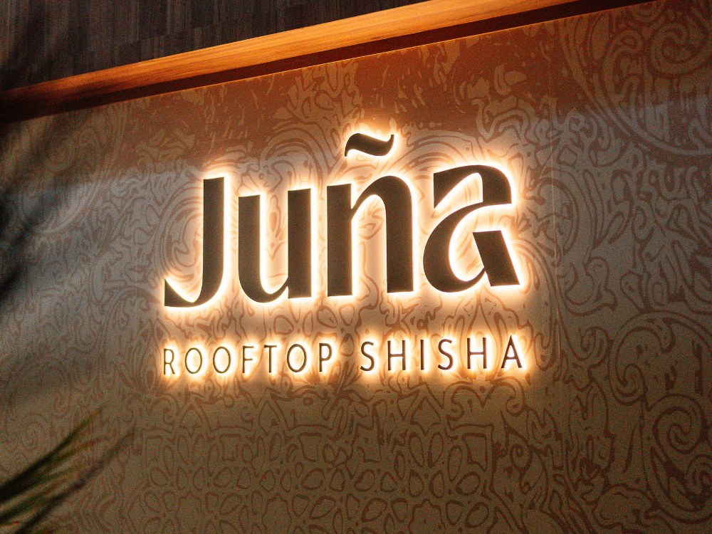 Illuminated sign for "Junā Rooftop Shisha" against a patterned wall, showcasing a vibrant and inviting atmosphere.