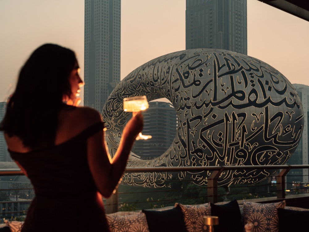 A woman raises a cocktail glass against the backdrop of a futuristic building adorned with Arabic calligraphy at sunset.