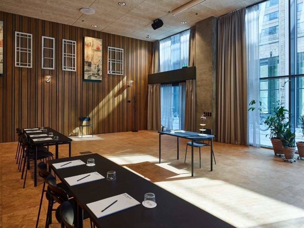 A modern conference room with wooden walls, tall windows, and sleek tables arranged in rows. Papers and glasses are neatly placed, creating a professional atmosphere. Sunlight streams in, adding warmth to the spacious, minimalist design.