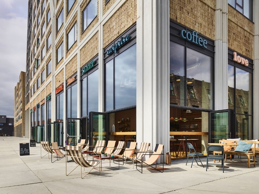 Hotel exterior with glass windows and open doors. Striped lounge chairs and tables are arranged outside on a sunny day, creating a relaxed atmosphere.
