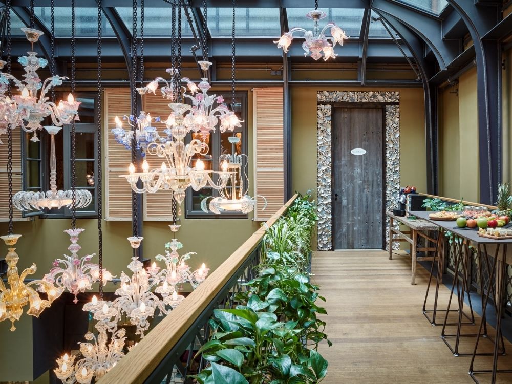 Brightly lit hallway featuring ornate chandeliers, lush plants, and a rustic door, with a table of fresh fruits in the foreground.