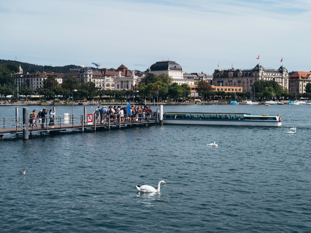 A busy dock by the lake in Zurich, with people boarding a boat and swans swimming nearby, set against a scenic cityscape.