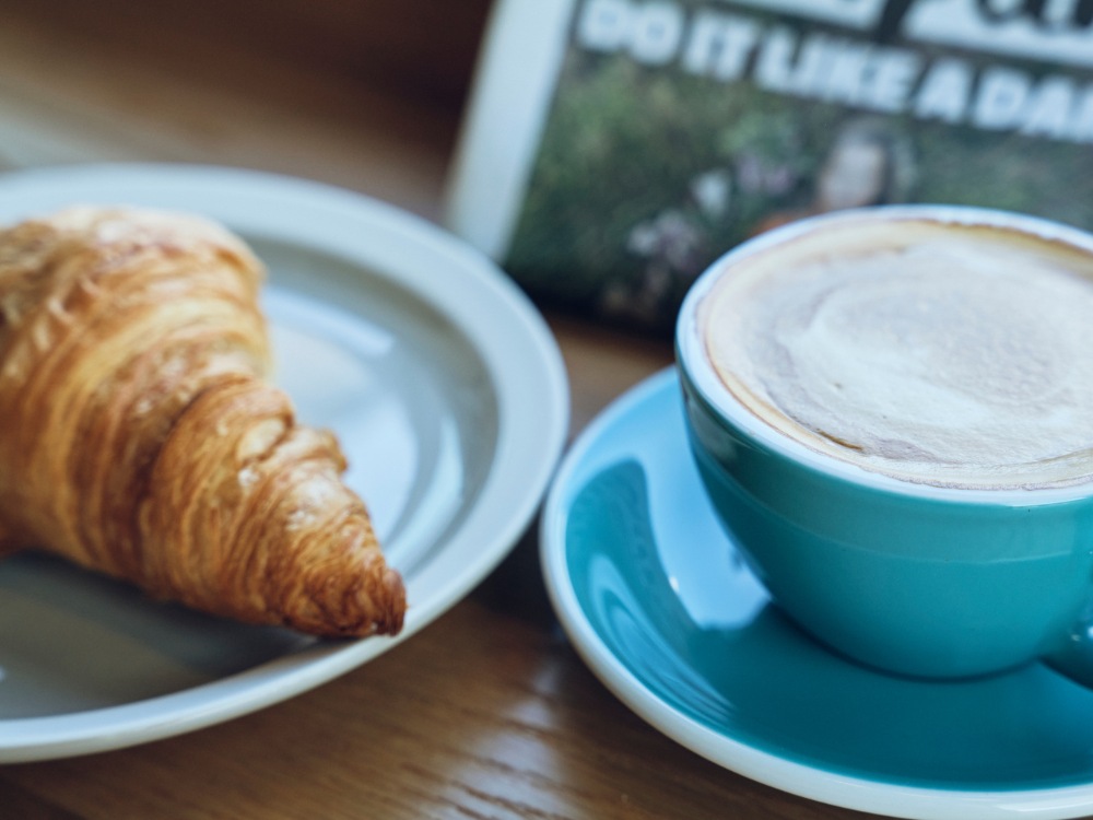 A croissant on a white plate sits next to a foamy cappuccino in a blue cup. The setting emits a cozy cafe atmosphere, suggesting a relaxed morning.