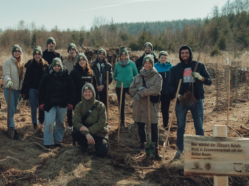 A group of people stands in a forested area, holding tools and posing beside a sign about a tree planting initiative.