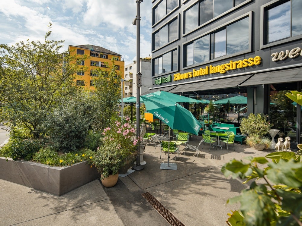 Urban hotel patio with vibrant green umbrellas and chairs, surrounded by lush plants. Modern building facade with large windows, sunny atmosphere.