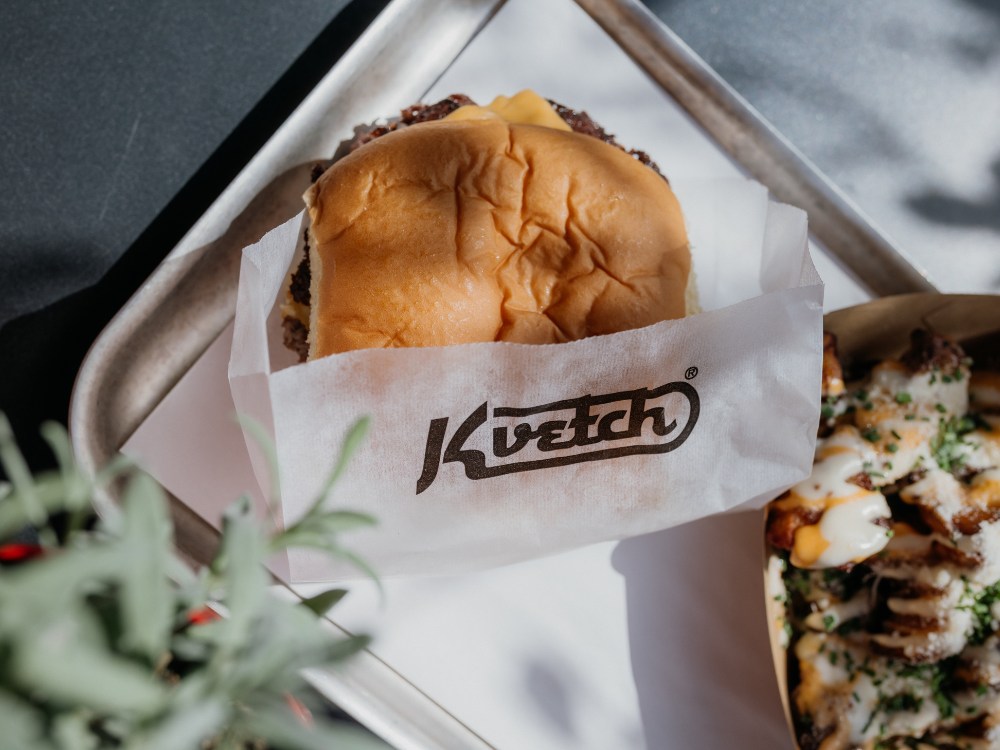 Burger wrapped in branded paper sits on a tray beside loaded fries, topped with melted cheese and herbs. Sunlight casts a warm, inviting glow.