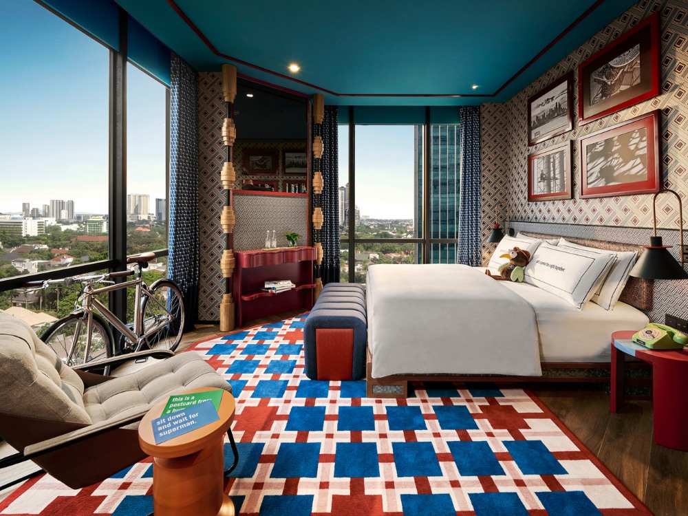 Hotel room featuring a colorful carpet, large windows with city views, a cosy bed, and stylish furnishings.