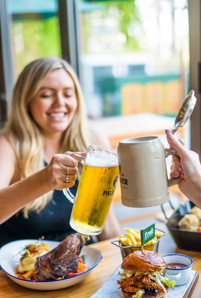 Two hands raise glasses in a toast, surrounded by hearty food items including a burger, fries, and a roasted meat plate.