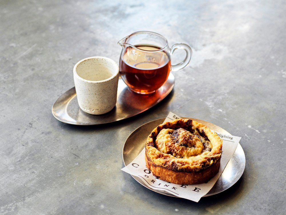 A rustic setting with a marble table holds a cup and pot of tea on a metal tray, paired with a cinnamon roll on a small plate on white paper.