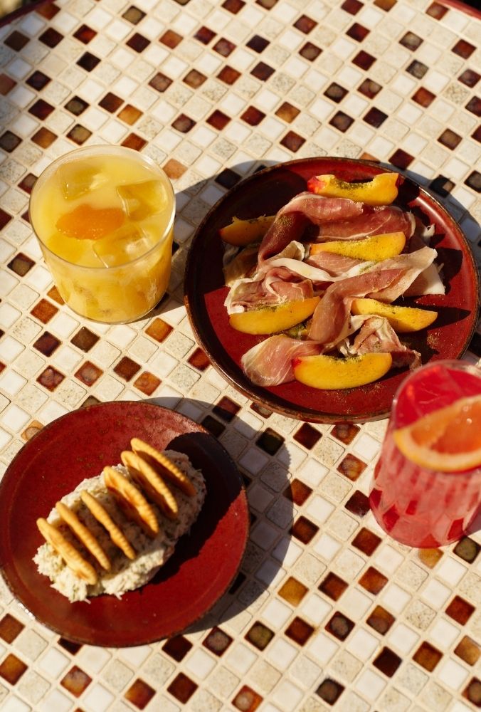 A sunlit table with a colorful mosaic pattern holds a red plate of sliced peaches and prosciutto. Nearby are crackers, dip, and two vibrant cocktails.