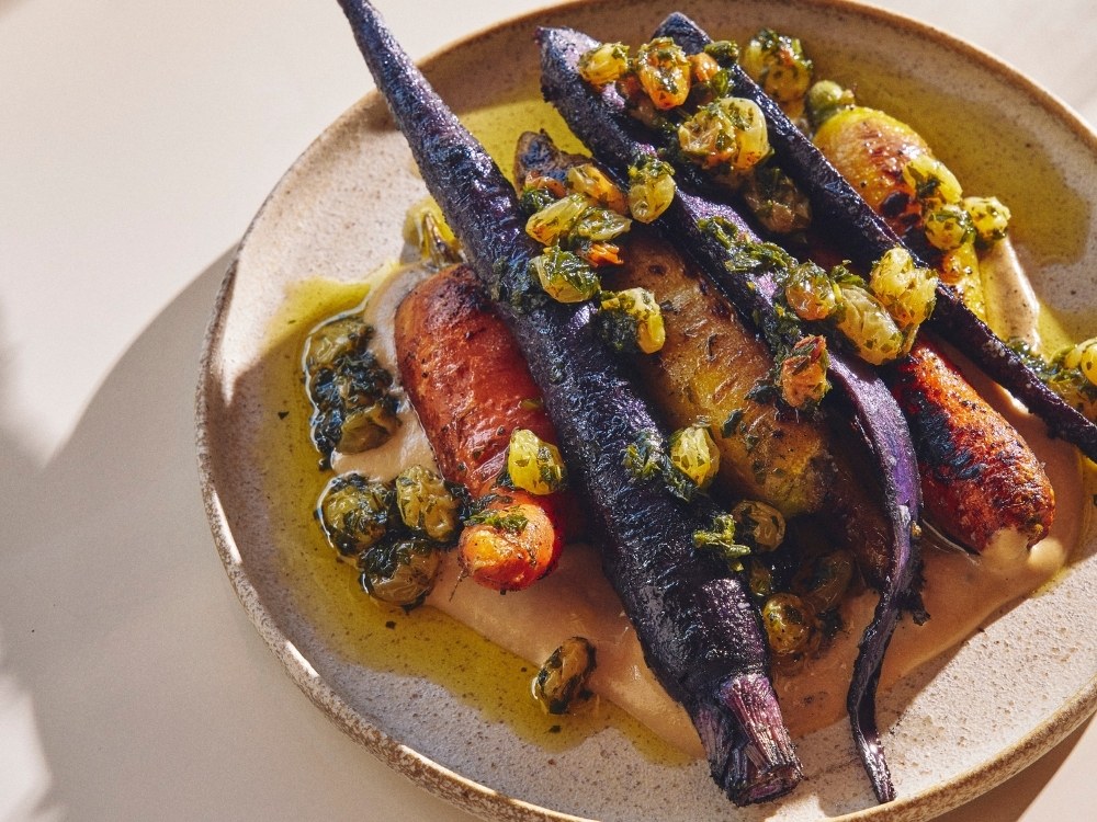 A plate of vibrant roasted carrots, both orange and purple, topped with golden raisins and herbs. Nestled on creamy puree, exuding a rustic, gourmet vibe.