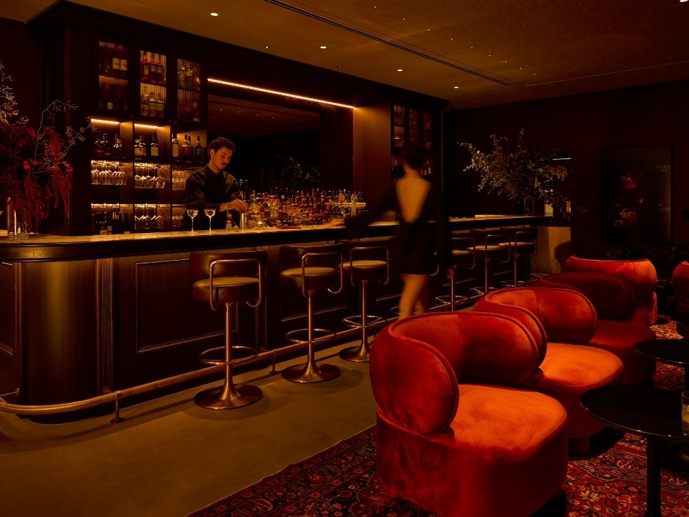 Sophisticated, dimly lit cocktail lounge featuring plush red velvet armchairs, an ornate oriental rug, a dark wood bar counter, and a bartender mixing drinks.