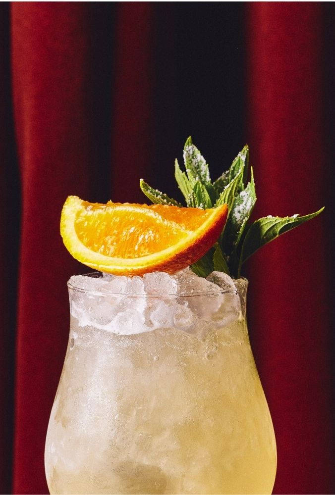 Chilled cocktail in a frosted glass garnished with an orange wedge and fresh mint, set against a rich, red curtain background, evoking elegance and refreshment.