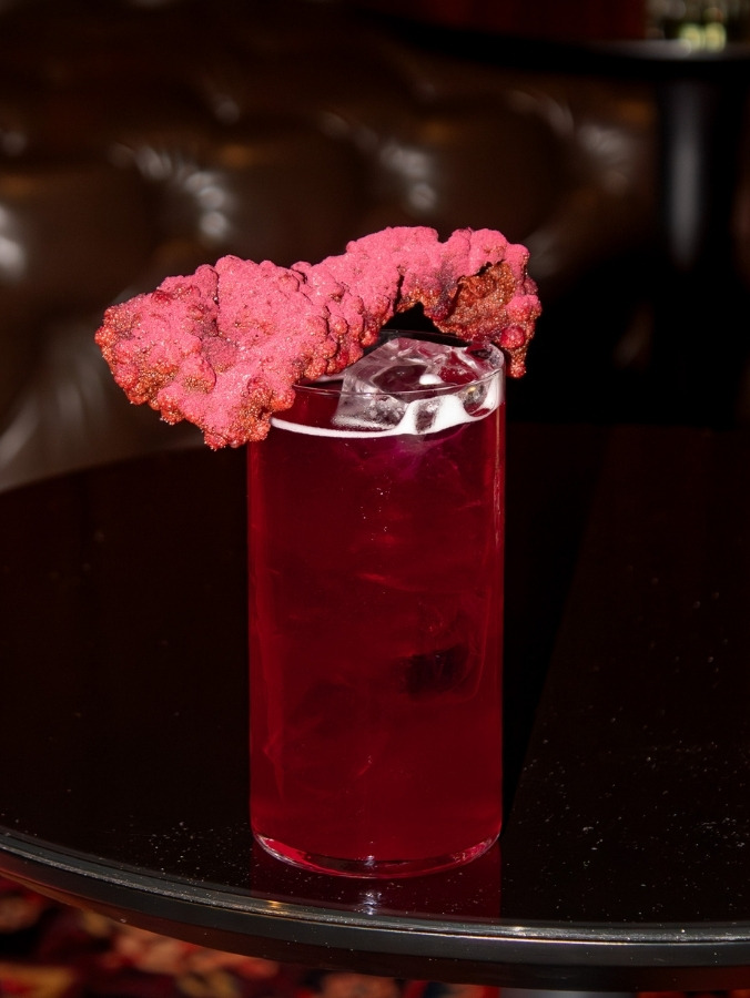 A vibrant red highball cocktail with ice, garnished with a unique textured pink coral-like crisp, sitting on a glossy black table.