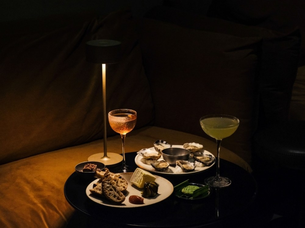 Moody hotel bar table setting featuring fresh oysters on ice, sliced sourdough bread with cheese, a slim gold cordless lamp, and two elegant coupe cocktails.