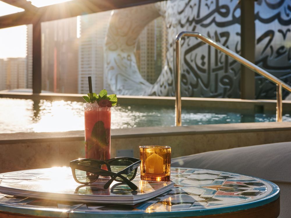A refreshing drink with mint and a candle on a mosaic table by a pool at sunset. Background features a modern building with intricate designs.