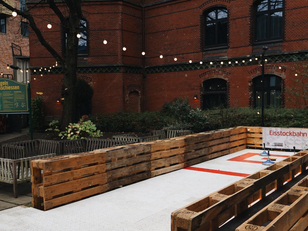 Outdoor ice rink surrounded by wooden barriers in a courtyard with string lights above, next to a brick building. A sign reads "Eisstockbahn".