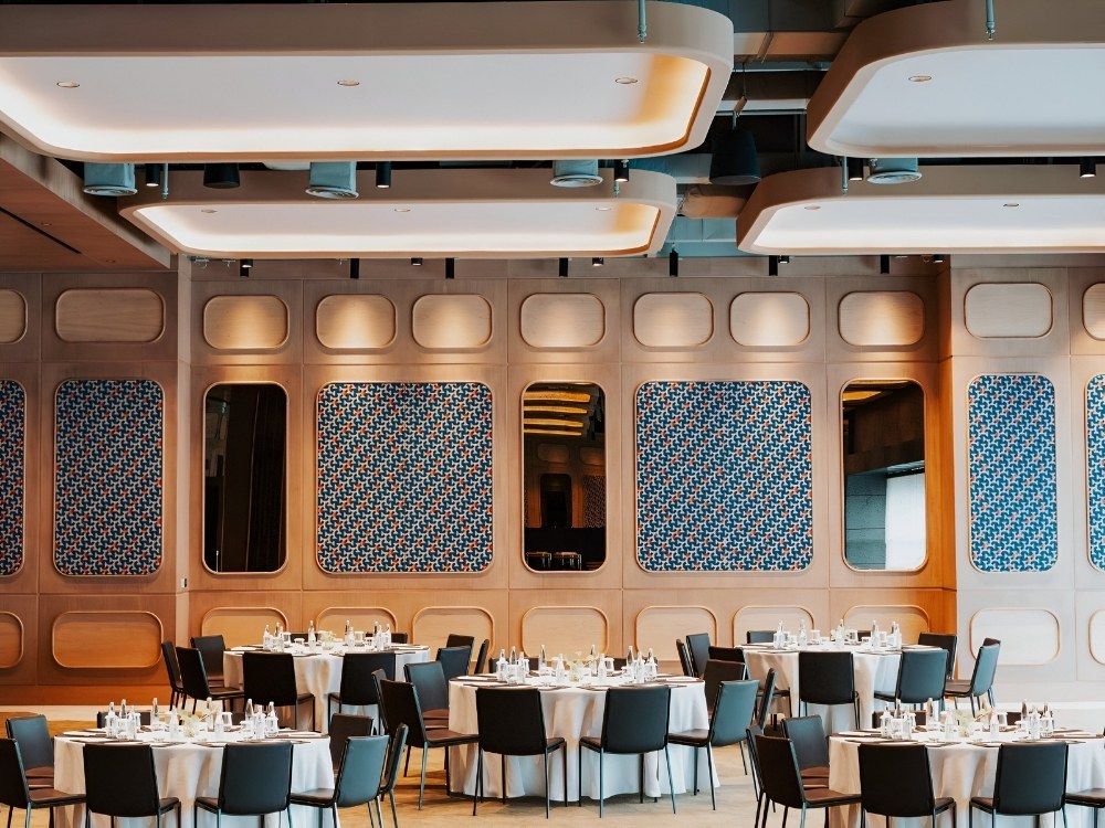 Event room with round tables set for dining, featuring a colorful patterned wall and modern, warm lighting.
