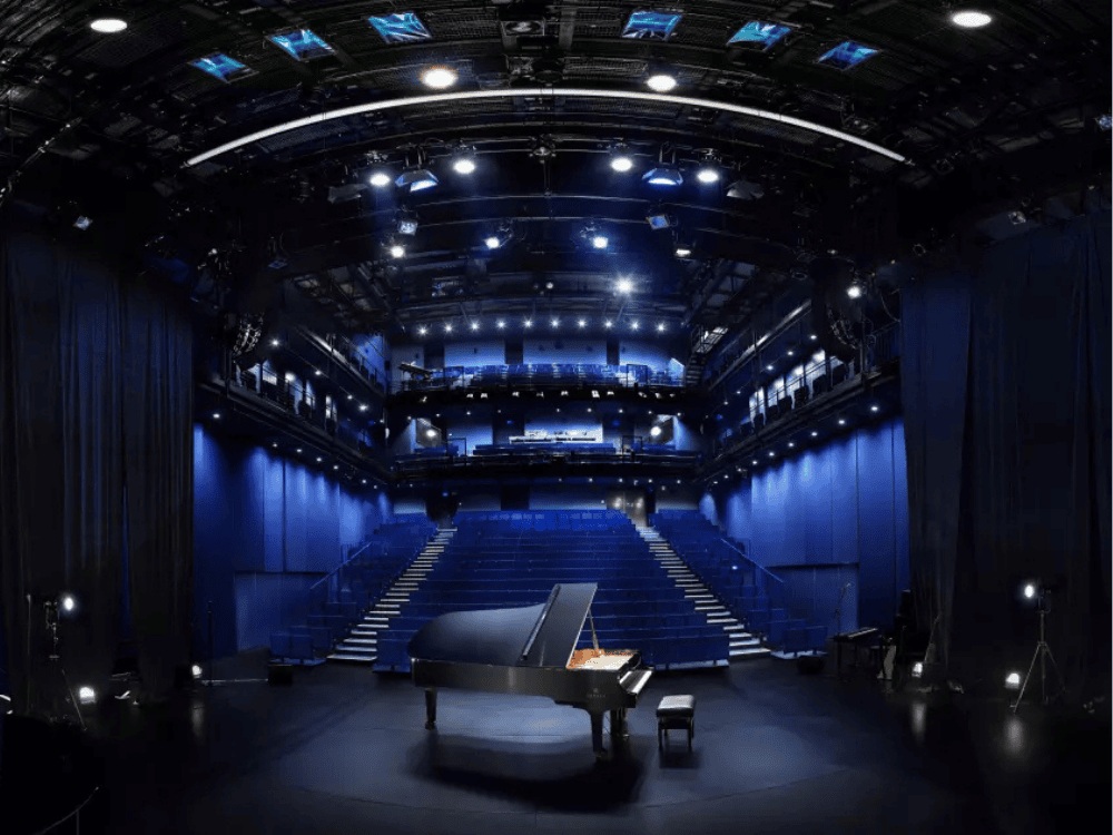 A grand piano on a dark stage, illuminated by blue lights, with tiered seating and lighting equipment visible in a modern theater setting.