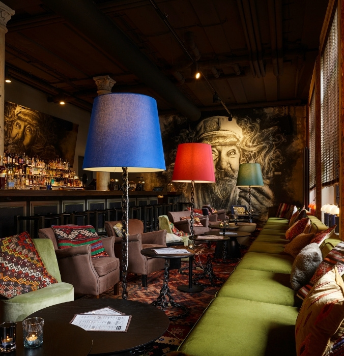 A cozy bar lounge with patterned cushions, round tables, colorful floor lamps, and large mural art of bearded face on the wall. Bottles line the shelves behind the bar counter.